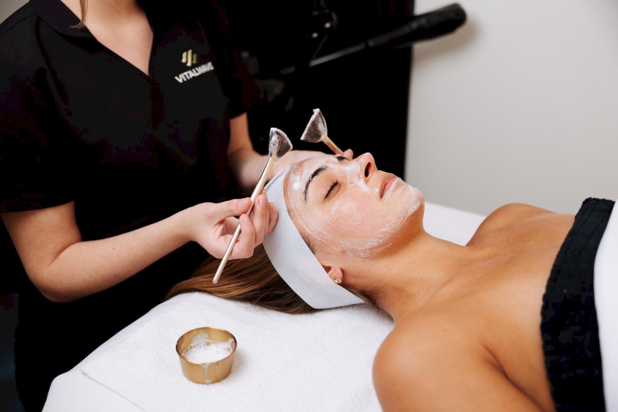 A woman lies with a facial mask applied while a skincare technician uses tools on her face; a small metal bowl sits nearby.