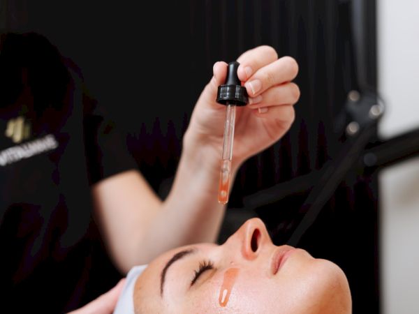 A person receives a facial treatment: serum drops applied with a dropper onto their forehead while a clinician shields and supports the head.