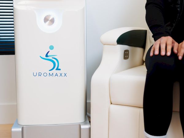 A medical emission device labeled &ldquo;UROMAXX&rdquo; sits beside a white chair; a person&rsquo;s legs and hands rest nearby.