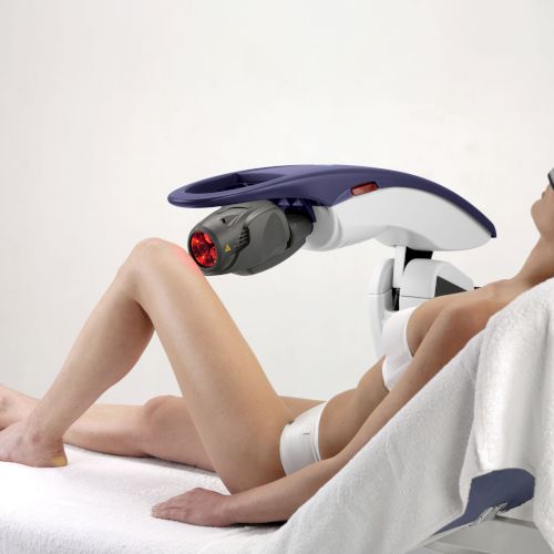 A person in protective eyewear undergoing a laser treatment while lying on a treatment table, with a large laser device positioned overhead.