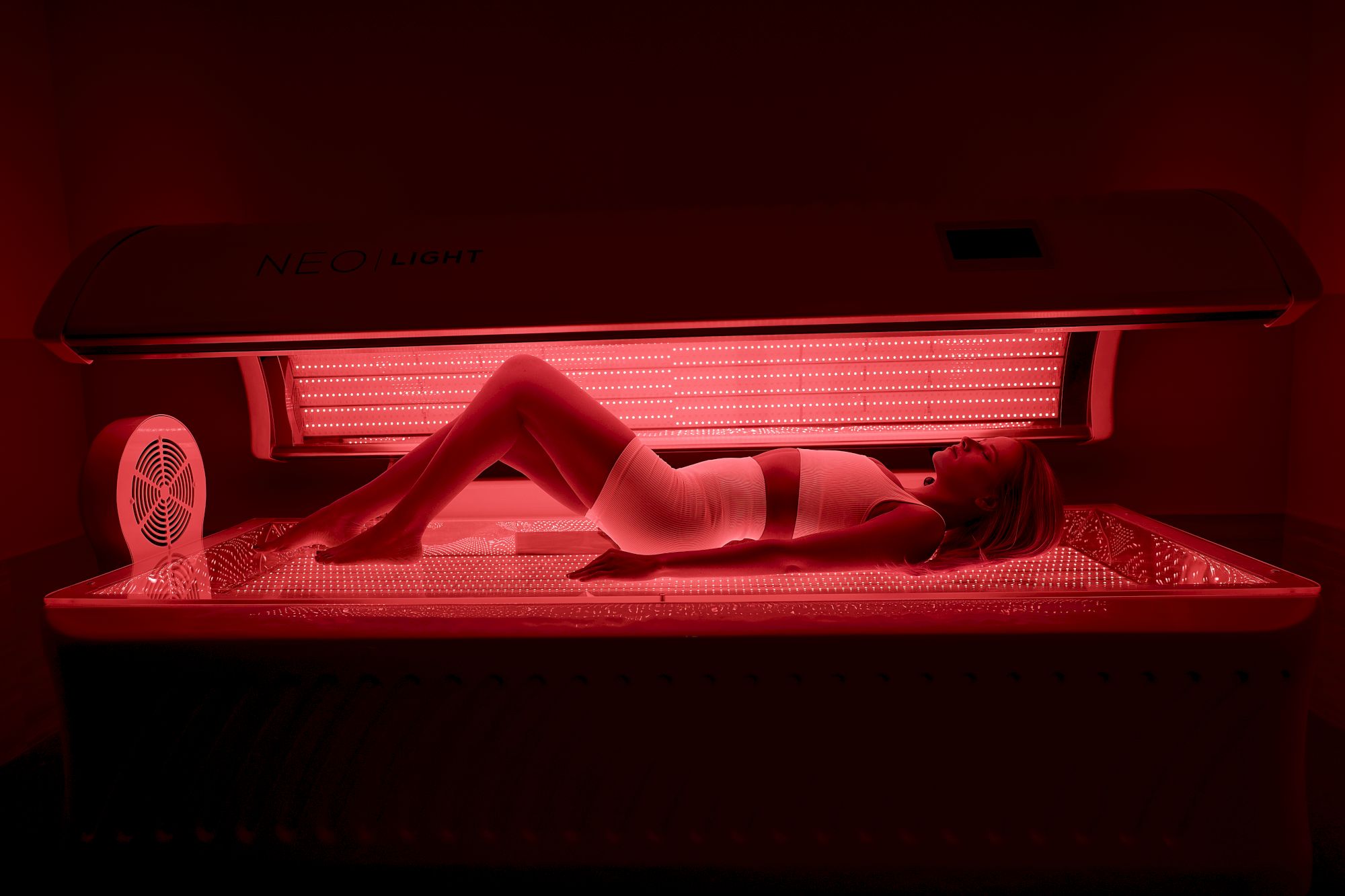 A person lies on a tanning bed under red UV lights, wearing minimal clothing, relaxing in a dimly lit room.