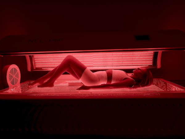 A person is lying inside a tanning bed, illuminated red by the UV lamps, wearing minimal clothing for tanning, with a fan visible on the side.