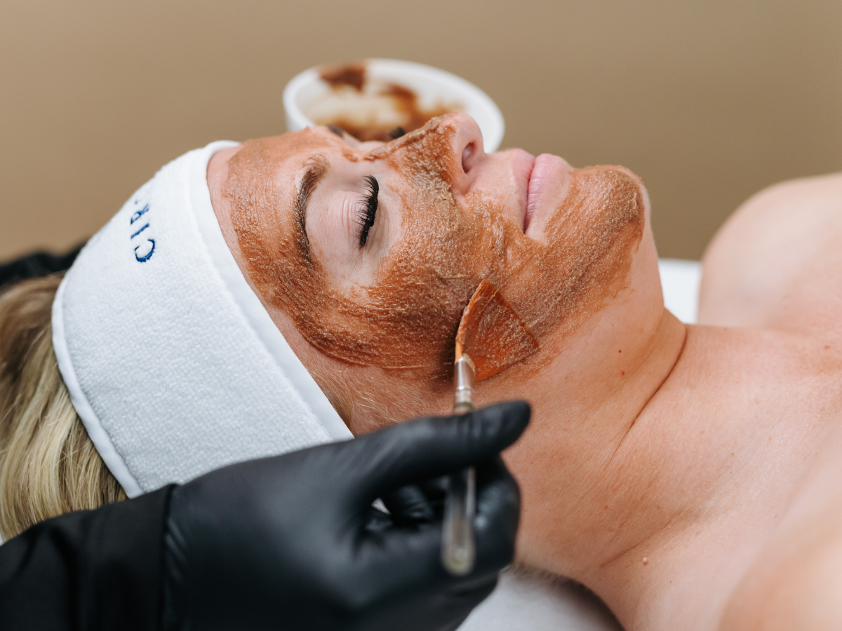A person is receiving a facial treatment with a brush applying a brown mask. They are wearing a headband, and hands are in black gloves.