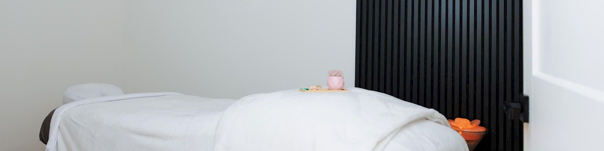 A minimalist spa-like room with a massage table covered in white sheets, a wall-mounted light fixture, and a tall black slatted divider next to a small wooden stool.