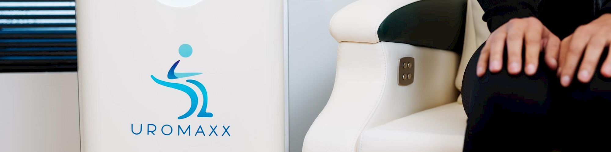 A medical device labeled &ldquo;UROMAXX&rdquo; beside a white chair; a person sits nearby with hands resting on their knee, in a clean, clinical room.
