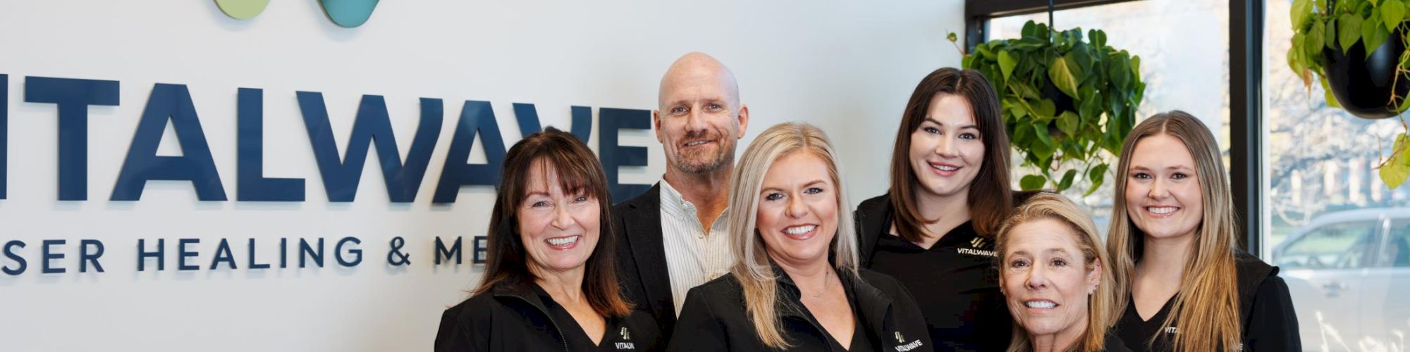 A group of six smiling professionals in black uniforms stands indoors in front of a wall with the logo &ldquo;NATALWA&hellip;&rdquo; and plants, posing for a team photo.