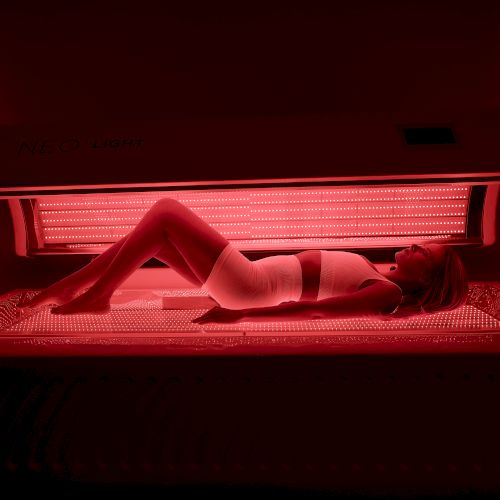 A person lies under a red tanning bed, wearing underwear, illuminated by the pinkish glow of the lamps.