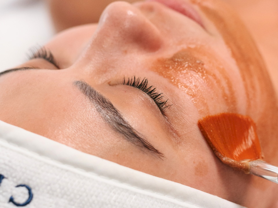 A person with a white towel wrapped head lying down, getting a facial treatment; a small brush applies a brown mask to the cheek area.