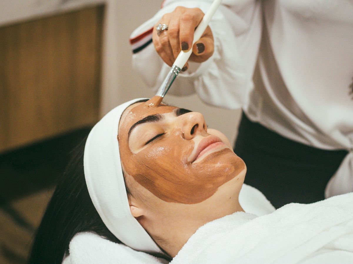 A person receives a facial treatment with a clay mask being spread on their face, lying down with eyes closed and head wrapped.
