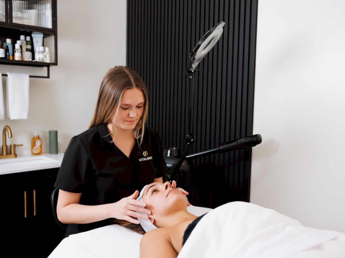 A skincare therapist in a black uniform gives a facial to a client lying on a treatment bed in a modern spa.