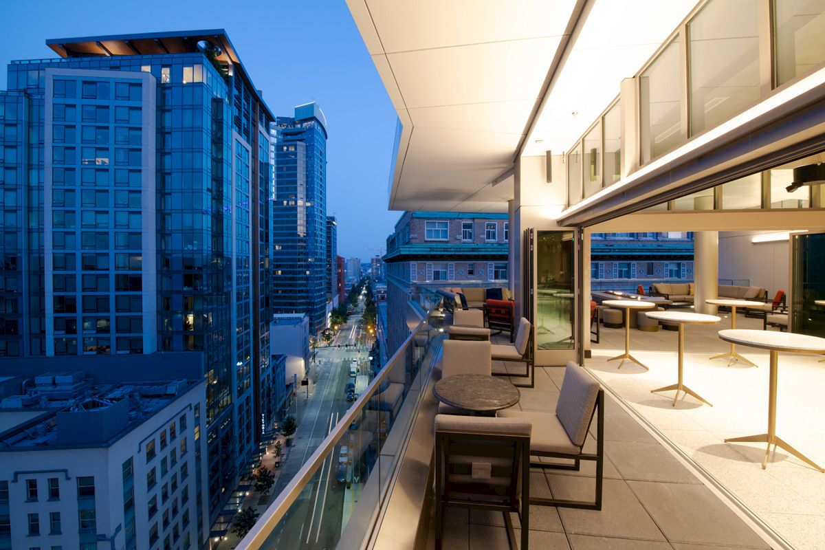 A modern rooftop terrace with seating overlooks a cityscape at dusk, featuring tall buildings and street views.