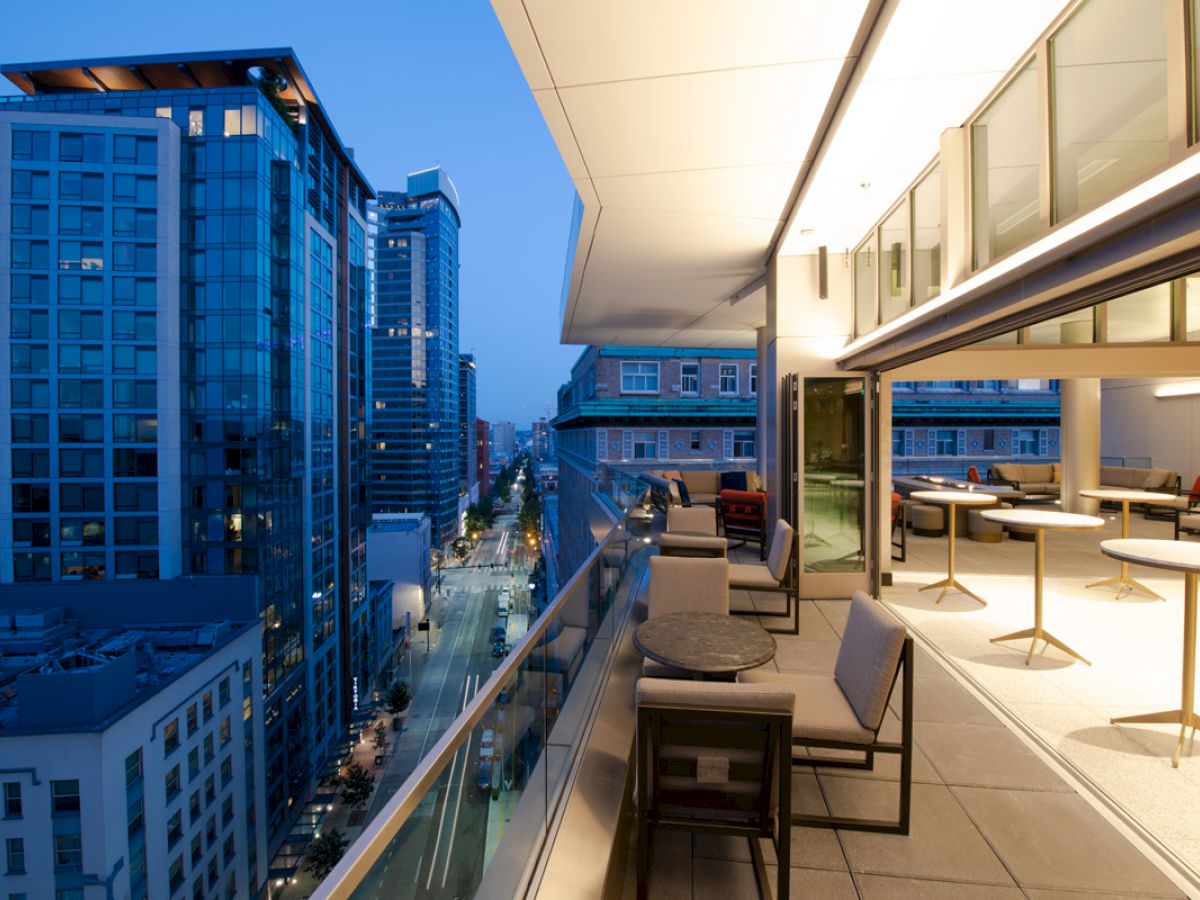 A modern rooftop terrace with seating and tables overlooks a city street lined with tall buildings at dusk.