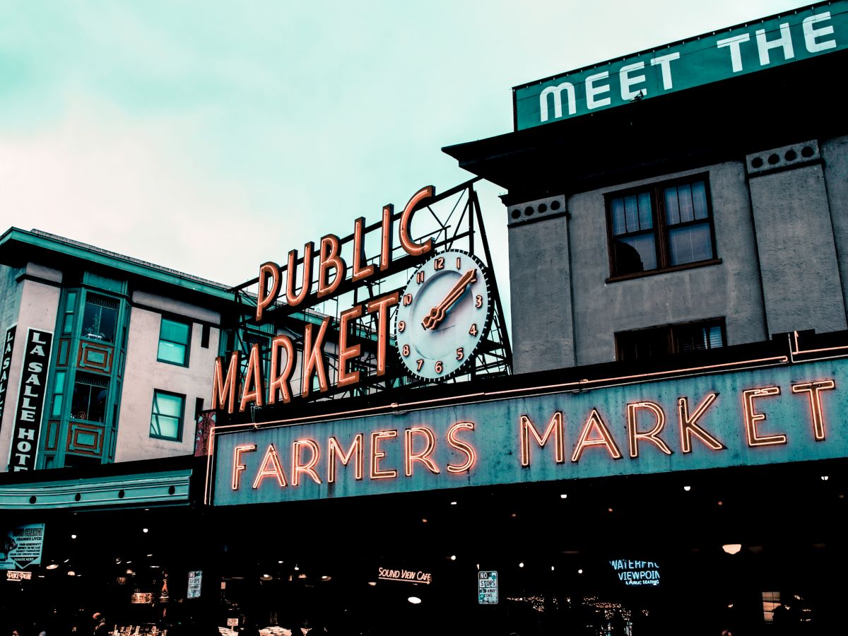 The image shows a neon sign for a public market and farmers market, with a large clock and surrounding buildings.