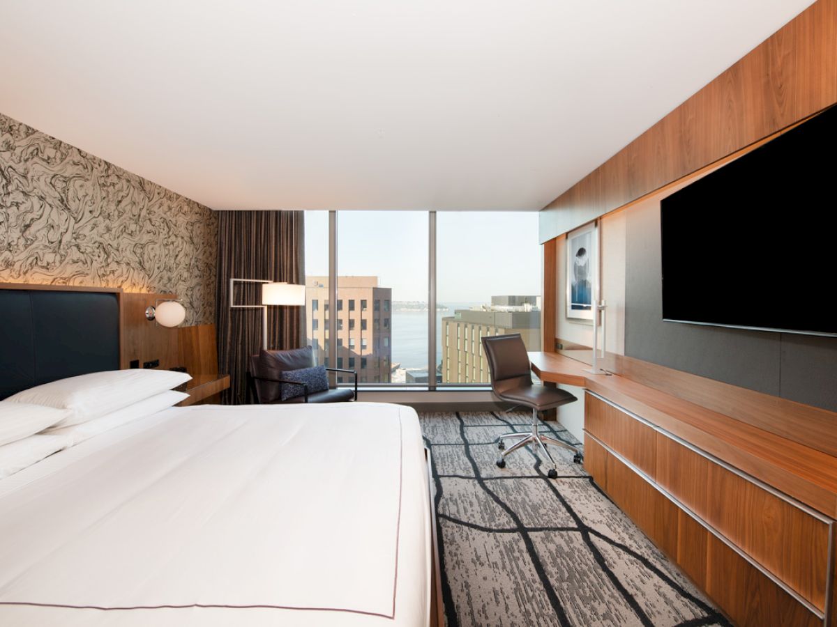 A modern hotel room with a large bed, a big TV, a desk, and a window offering a city view is shown in the image.
