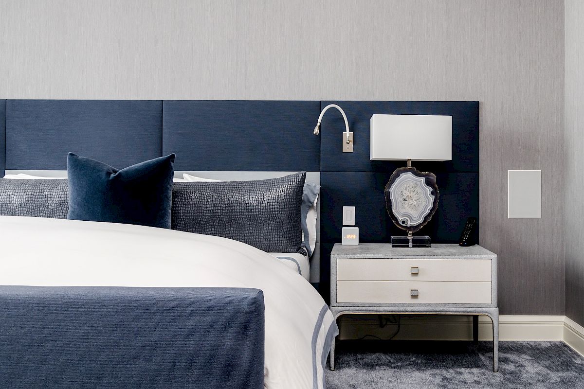 A modern bedroom with a blue and white color scheme, featuring a bed, pillow, a nightstand with a lamp, and decorative items on a carpeted floor.